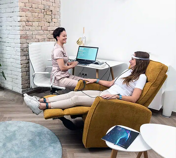 a woman sitting in a yellow reclining chair connected to equipment while a therapist works on a computer in a modern office promoting relaxation and wellness along with eight key benefits of therapy